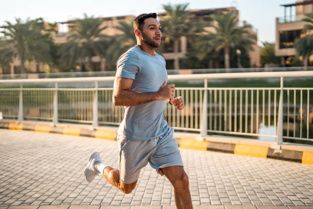 A mid-adult Hispanic man in athletic attire runs outdoors on a paved pathway in Dubai at sunrise, surrounded by palm trees and modern buildings.